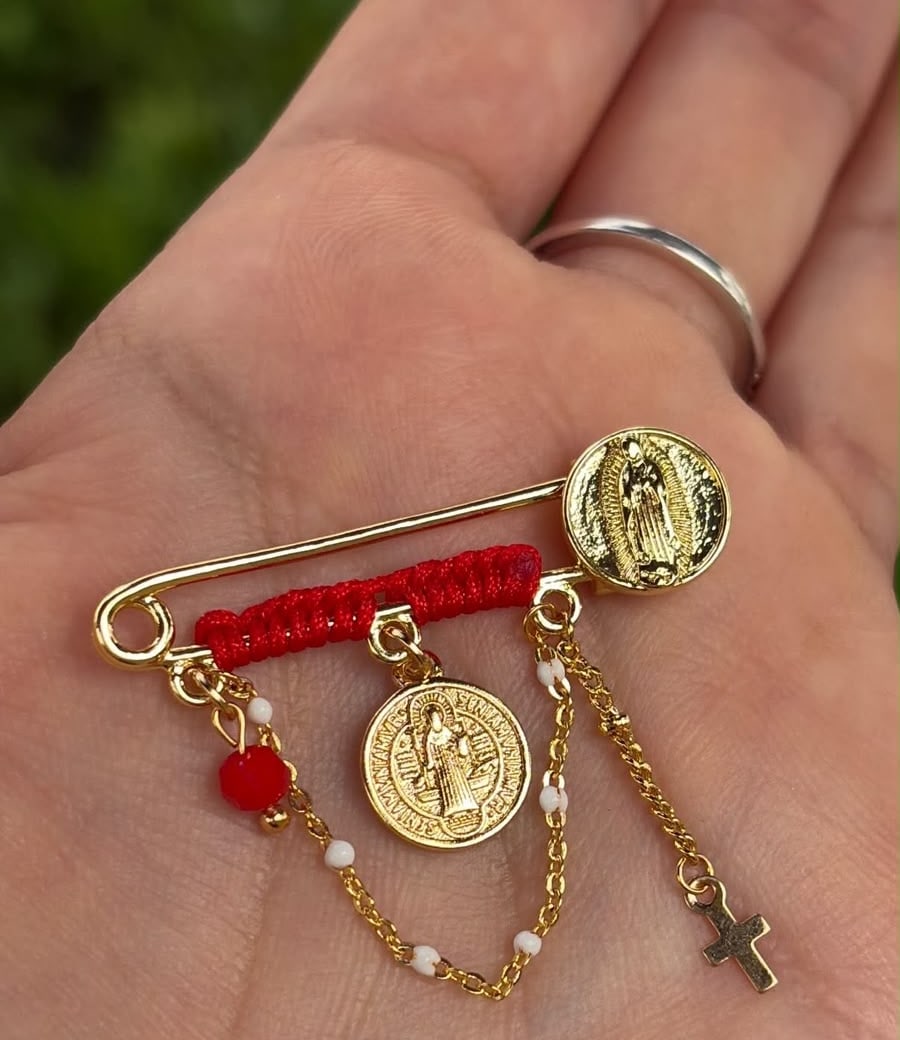 Prendedor con hilo rojo, corazón rojo, medallas, cadena y cruz.