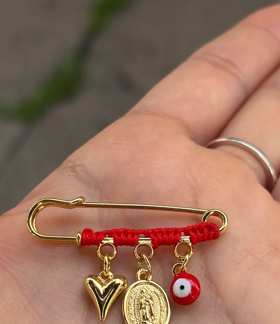 Prendedor con hilo rojo, corazón, medalla y ojo rojo, sobre la palma de una mano.