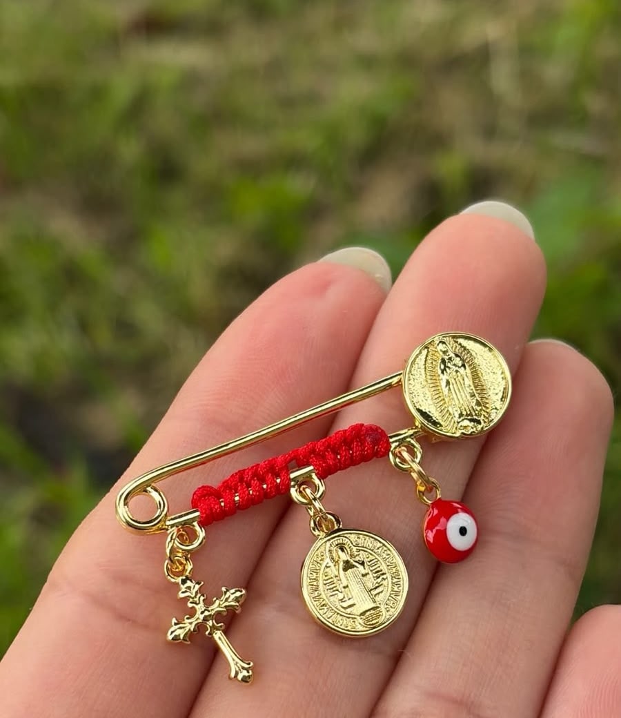 Prendedor con hilo rojo, cruz, medallas y ojo rojo.