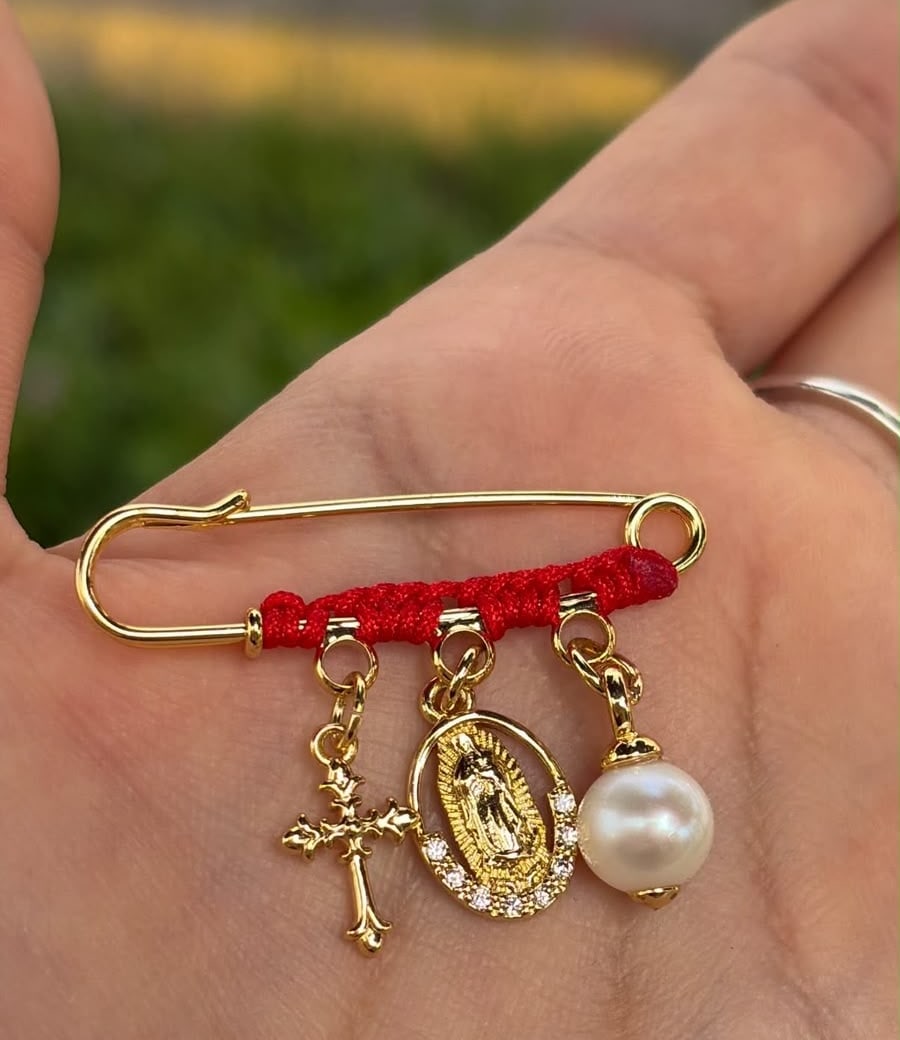 Prendedor con hilo rojo, cruz, medalla de la Virgen y perla.