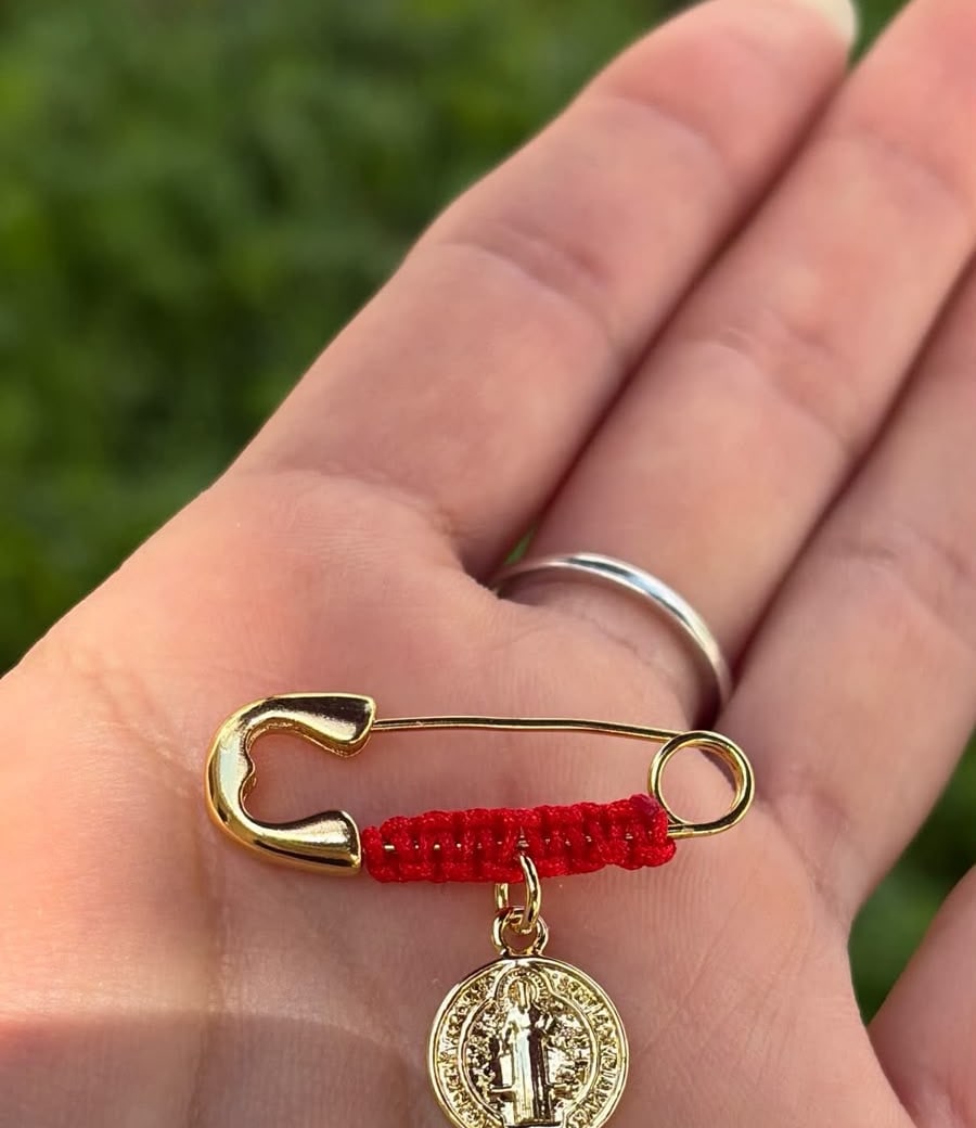 Prendedor con hilo rojo y medalla de San Benito, sobre la palma de una mano.